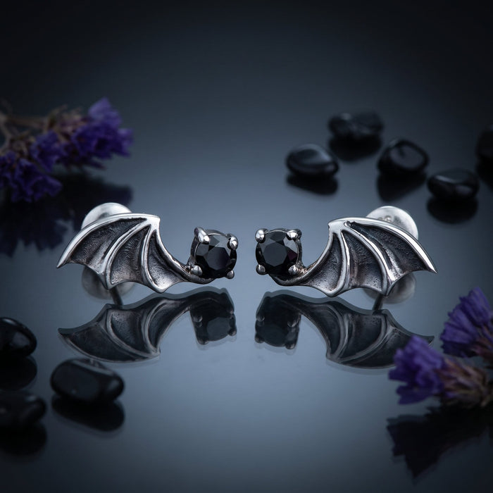 Moonlit still life of both black cubic zirconia bat earrings on smoked glass with obsidian pebbles