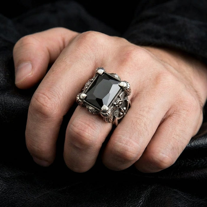 Men's silver dragon claw ring with large rectangular black onyx gemstone worn on middle finger, featuring gothic ornate detailing and prong setting against black leather background