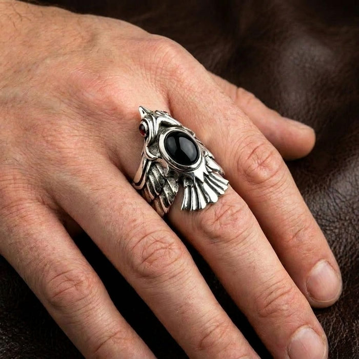 Close-up of a man's hand wearing a detailed silver phoenix biker ring featuring a prominent oval black onyx stone and a red gemstone eye, resting against a textured brown leather background.