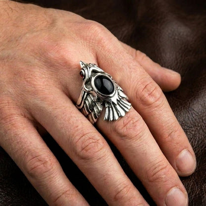 Close-up of a man's hand wearing a detailed silver phoenix biker ring featuring a prominent oval black onyx stone and a red gemstone eye, resting against a textured brown leather background.
