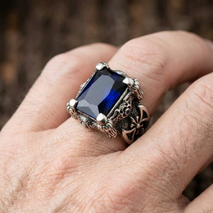 Large rectangular blue sapphire ring with silver dragon claw prongs and gothic cross detailing worn on a man's finger, showcasing intricate biker-style metalwork and dark oxidized finish