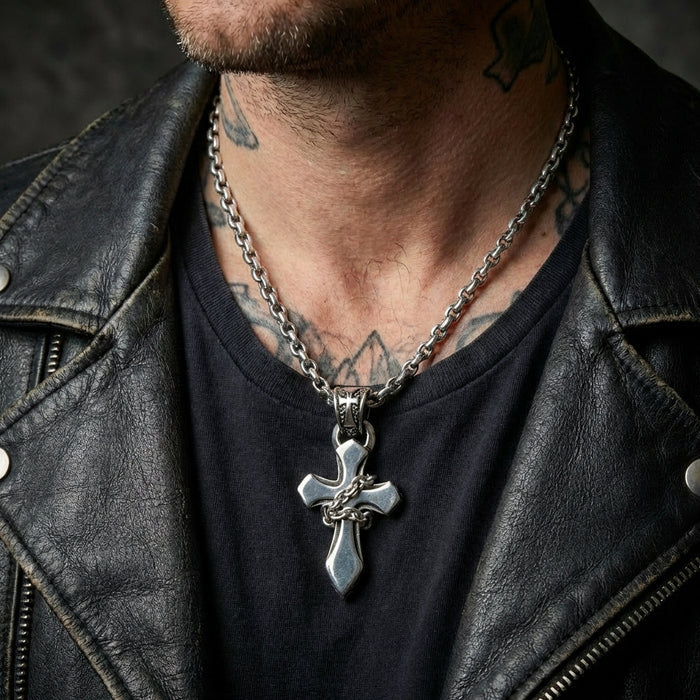 Close-up of a male model wearing a sterling silver chain-wrapped gothic cross pendant on a thick link chain, styled with a distressed black leather biker jacket and visible neck tattoos.