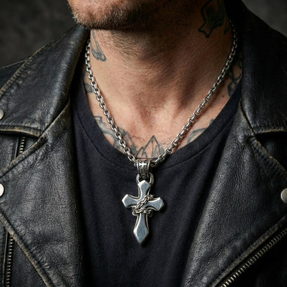 Close-up of a male model wearing a sterling silver chain-wrapped gothic cross pendant on a thick link chain, styled with a distressed black leather biker jacket and visible neck tattoos.