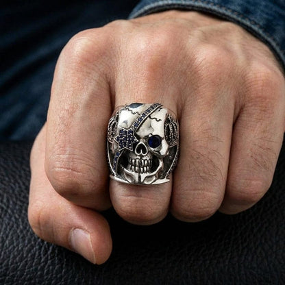 Sterling silver crown skull ring with blue sapphire eye patch and pavé stones worn on a man's middle finger against a dark leather and denim background