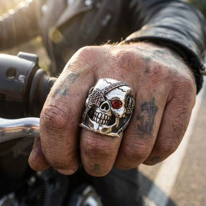 Tattooed biker hand gripping a motorcycle handlebar wearing a large sterling silver skull ring featuring a red gemstone eye and a studded star eye patch.