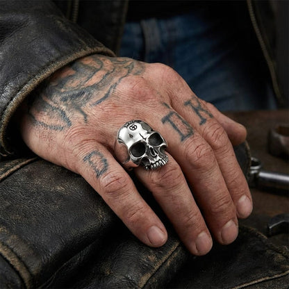 Close-up lifestyle shot of a man's tattooed hand resting on a distressed black leather jacket, showcasing a heavy sterling silver half-jaw skull biker ring.