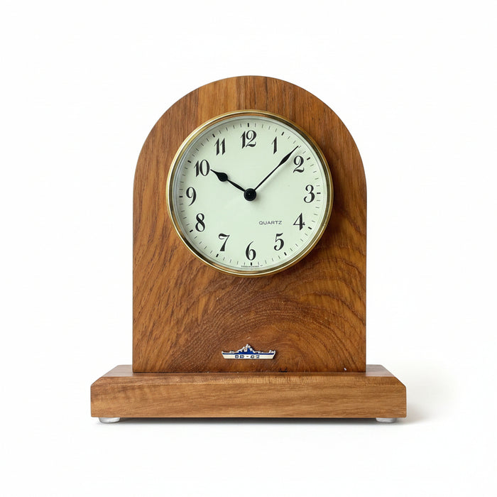 A vintage-style wooden desktop clock with an arched top and a small base, featuring a cream-colored dial with black numerals and hands. The clock is photographed on a dark desk surface next to a calculator.