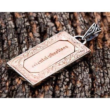 Recessed copper plaque inside an engraved silver border, the openwork bail above it, laid on dark wood.