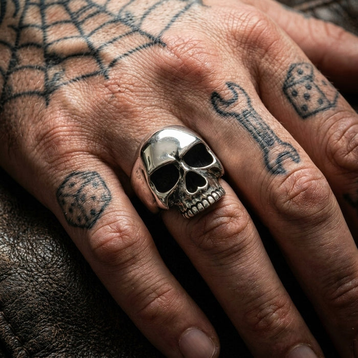 Keith Richards sterling silver skull ring worn on the middle finger of a tattooed biker hand with spider web, dice, and wrench tattoos on dark leather.