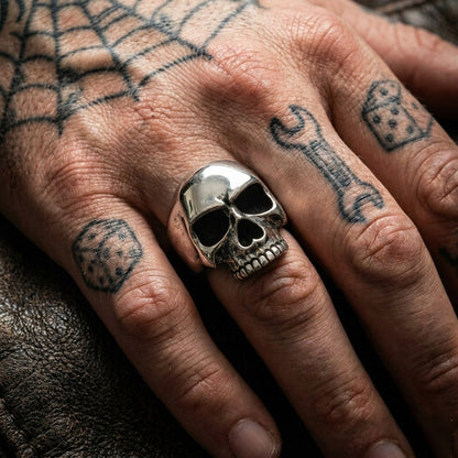 Keith Richards sterling silver skull ring worn on the middle finger of a tattooed biker hand with spider web, dice, and wrench tattoos on dark leather.