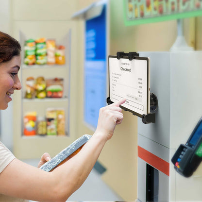 A woman uses a self-checkout kiosk in a grocery store, interacting with a touchscreen POS Tablet securely positioned on the sleek LockVise™ POS Drill-Base Locking Tablet Mount to complete her purchase while holding a food tray. Shelves with packaged goods are visible in the background.
