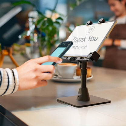 A person holds a phone near a tablet on the LockVise™ POS Locking Tablet Stand at a counter, making a contactless payment. The tablet screen shows “Thank You” and “PAID $11.00,” with a coffee cup and plant in the background.