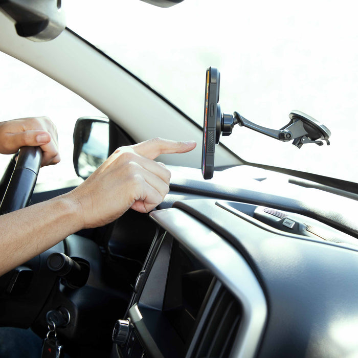 While steering with one hand, a person reaches for their smartphone held in a Magnetic Phone Holder with Sticky Suction Mount - MagSafe Compatible, securely attached to the dashboard.