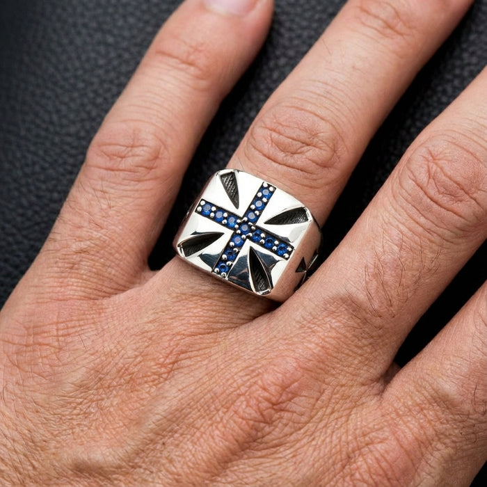 Silver Maltese cross ring with blue gemstones worn on middle finger against dark textured background showing intricate cross design and pavé setting