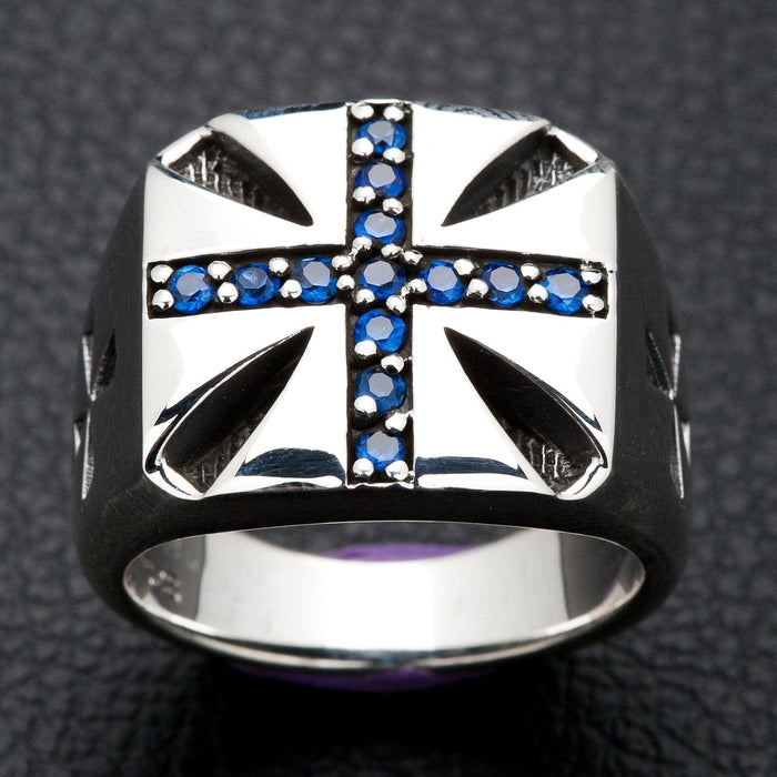 A solid sterling silver Maltese cross ring, inlaid with brilliant blue stones, shown on a dark, textured background.