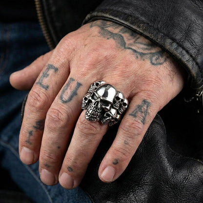 A close-up lifestyle shot of a polished silver phantom skull ring featuring multiple skulls, worn on the middle finger of a man's tattooed hand resting against a black leather biker jacket.