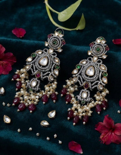 A pair of ornate Indian chandelier earrings with silver-toned metalwork, white faux pearls, and red and green stones, displayed on a dark green velvet fabric with scattered flower petals.