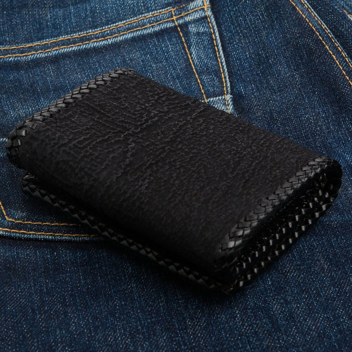 Woven leather lacing edges the black shark hide, the closed wallet resting on blue denim.