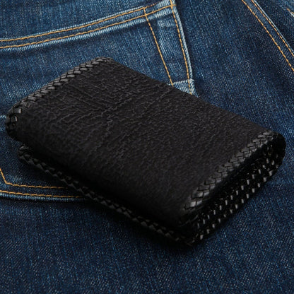 Woven leather lacing edges the black shark hide, the closed wallet resting on blue denim.