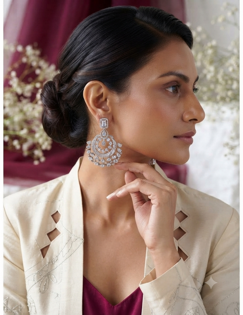 A woman in a cream-colored embroidered jacket and maroon top wears a pair of ornate silver-toned statement earrings with clear stones and orange stone accents, posed against a soft-focus background of burgundy drapery and white flowers.