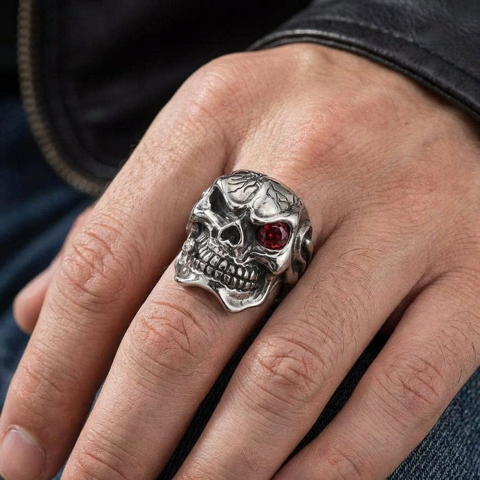 Silver cracked skull ring with red gemstone eye worn on man's hand, featuring detailed teeth and textured design, styled with leather jacket and denim jeans