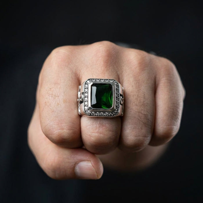 Close-up of a man's fist wearing a bold sterling silver gothic ring featuring a large rectangular green gemstone surrounded by a crystal halo against a black background.