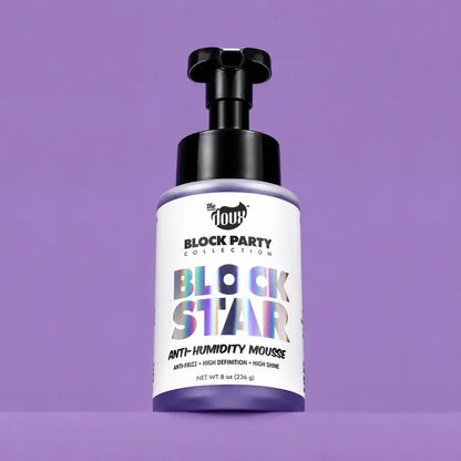 A bottle of The Doux Block Party Collection Block Star Anti-Humidity Mousse, featuring a white label with holographic lettering and a black pump dispenser, set against a solid purple background.